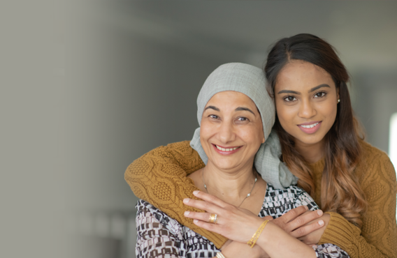 A younger woman hugging an older woman from behind. Both are smiling and the older woman has a cloth wrapped on top of her head.