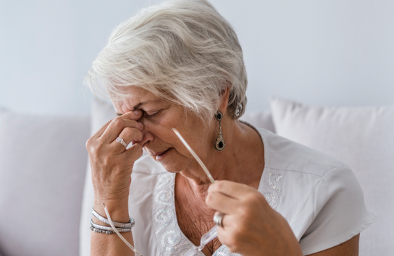 An older woman with gray hair taking off her glasses and holding her nose grimacing.