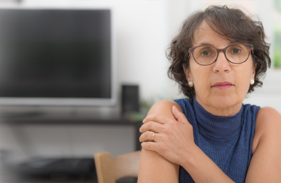 A brunette woman wearing glasses in front of a computer holding her shoulder.