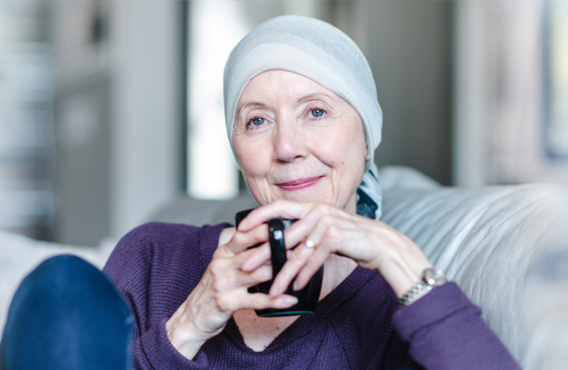 Smiling woman with a cloth wrapped on top of her head sitting on a couch with a coffee mug