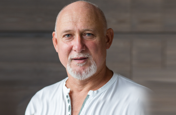 A older bald man standing in front of a blurred wooden wall.