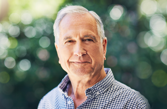 Smiling old man with gray hair standing outside in front of some bushes.