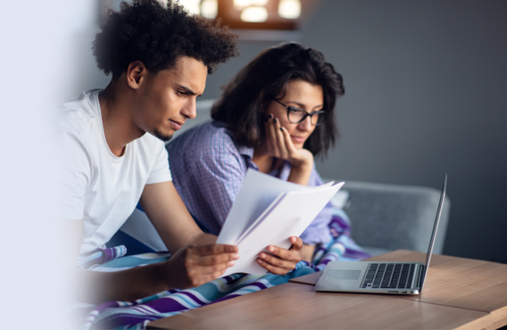 A man and woman sitting on a couch. The man is reviewing papers on a table and the woman is looking at a laptop on the table.