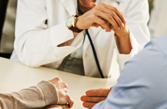 An image of two people talking with a doctor. The view point is behind the two people with a focus their hands on a table and the doctor's hands closed together.