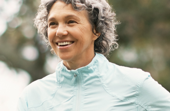 Smiling person with curly hair wearing a light blue jacket, standing in front of a blurred background with trees. To the right, a red blood drop icon with a white heart is displayed on a pink background.