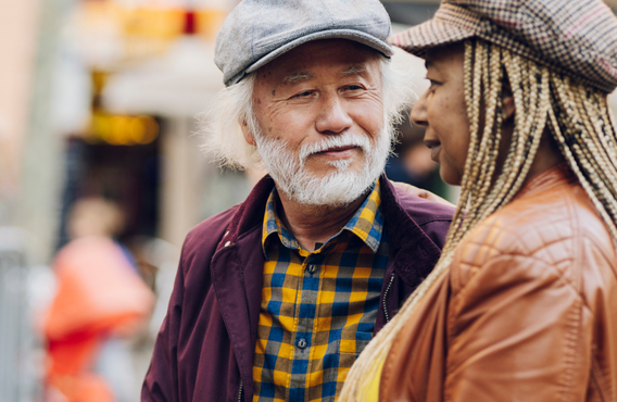 An older Asian man with long hair looks lovingly at a Black woman with braids. They are on a city street.
