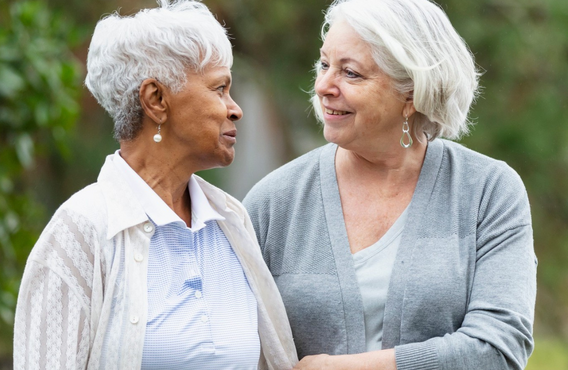 Two older adults walking outdoors side by side, looking at each other and smiling, with greenery in the background.