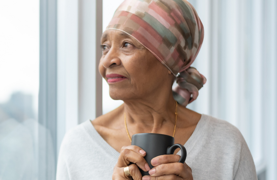 Person wearing a patterned headscarf and holding a mug while standing beside a large window.  Soft natural light illuminates the scene, with a blurred city skyline visible in the background.