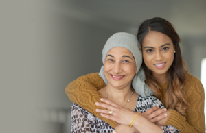 A younger woman hugging an older woman from behind. Both are smiling and the older woman has a cloth wrapped on top of her head.