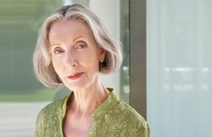 A woman with blonde-gray hair standing by a window that is blurred in the background.