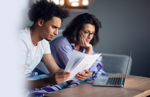 A man and woman sitting on a couch. The man is reviewing papers on a table and the woman is looking at a laptop on the table.