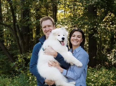 Steve Hopkin and Shannon holding a white dog outside in front of a sunny wooded area.