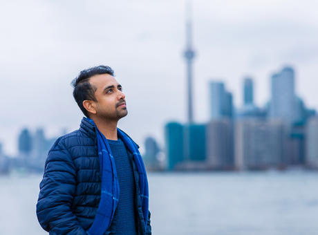 A man standing by a harbor in Toronto staring off into the distance with his hands in his pockets. Behind him is the landscape of downtown Toronto with the CN tower visibile.