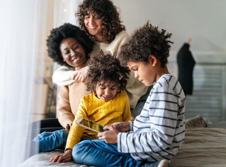a family of 4 sitting on a bed. The two parents are hugging and the two of the kids are playing with a tablet.