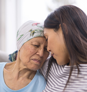 An elderly woman with myeloma wearing a head cloth hugging and laying her head against another woman.