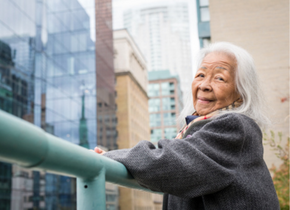A woman with long white hair stands outside on a balcony in a city and looks at the camera