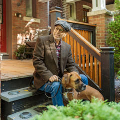 A man dressed in a tweed jacket and hat sits on a stoop. His dog is with him.