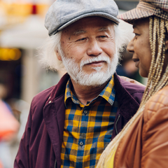 An older Asian man with long hair looks lovingly at a Black woman with braids. They are on a city street.
