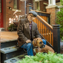 A man dressed in a tweed jacket and hat sits on a stoop. His dog is with him.