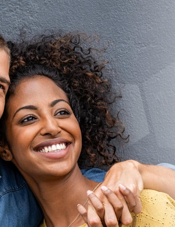 A man hugging a woman, both smiling with their backs against a wall.