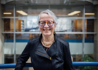 Camille, an elderly white woman with gray hair and glasses smiling. She is inside a building leaning on a railing with interior windows far behind.