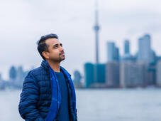 A man standing by a harbor in Toronto staring off into the distance with his hands in his pockets. Behind him is the landscape of downtown Toronto with the CN tower visibile.