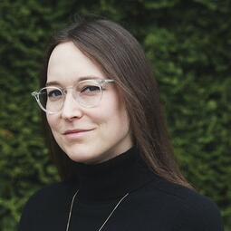 Une femme portant des lunettes est habillée en noir et regarde la caméra. Elle se tient devant un mur de verdure.