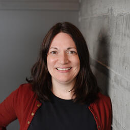 A woman with a red cardigan stands against an exterior wall. She is smiling.