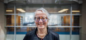 Camille, an elderly woman with gray hair and glasses smiling. She is inside a building leaning on a railing with interior windows far behind.