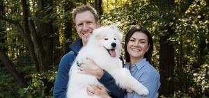 Steve Hopkin and Shannon holding a white dog outside in front of a sunny wooded area.