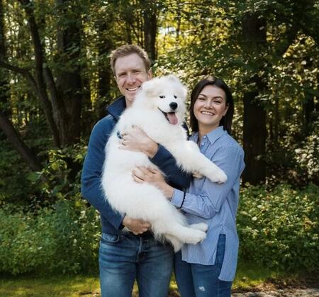 Steve Hopkin and Shannon holding a white dog outside in front of a sunny wooded area.