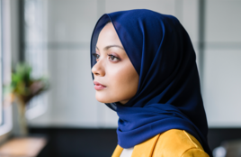 Woman wearing blue hijab standing by a window looking outside.