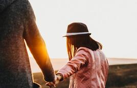 Person in a pink jacket and hat walking across an open landscape while holding another person’s hand.  Warm sunset light shines between them, creating a bright glow against the natural background.